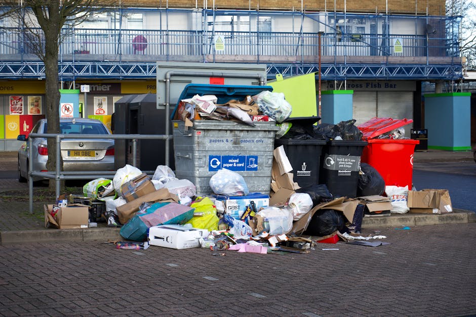 A large green outdoor waste bin with the number 53 and the words 'JUN BESAR' printed on it, positioned on a concrete sidewalk in front of a metal shelving unit holding cleaning supplies and tools. The
