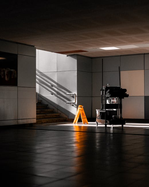 An indoor stairwell with stainless steel handrails and tiled walls, illuminated by overhead lighting that casts shadows across the tiled floor. A bright orange caution sign indicating a wet surface is